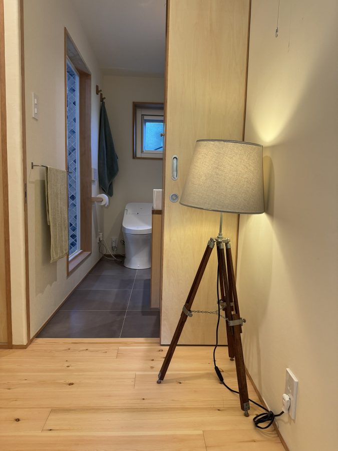 三脚のフロアランプを灯すと、やわらかな光が壁に広がり、木の床や建具の温かさをより引き立ててくれます。 奥に見える洗面やトイレ空間とのつながりも、暮らしの一部として自然に溶け込み、ほっと落ち着ける雰囲気に。 シンプルな中に、ちょっとしたデザインの工夫で空間が豊かになる瞬間です。